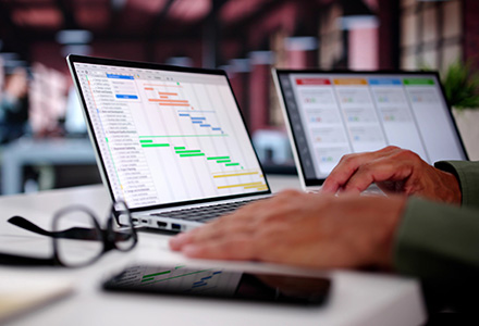 Two laptop screens with a Gantt chart and project management tool on a desk in an office