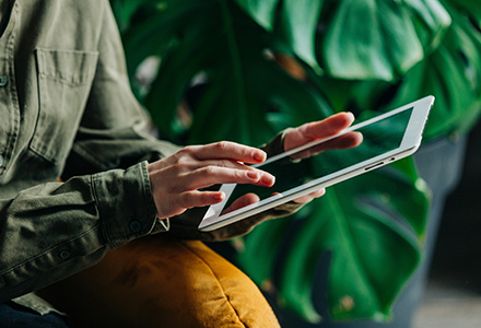 Woman scrolling on a tablet with a large plant behind her