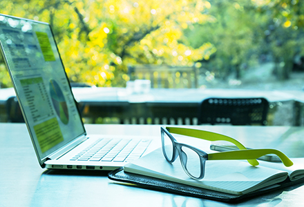 Photo of a laptop on a table with papers in front of it and a pair of glasses on top