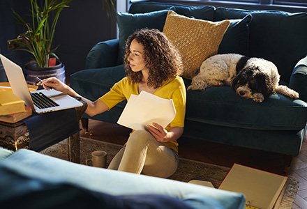 Woman at home next to couch on laptop with dog in background