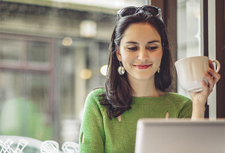 woman looking at a laptop, holding a coffee mug, and smiling